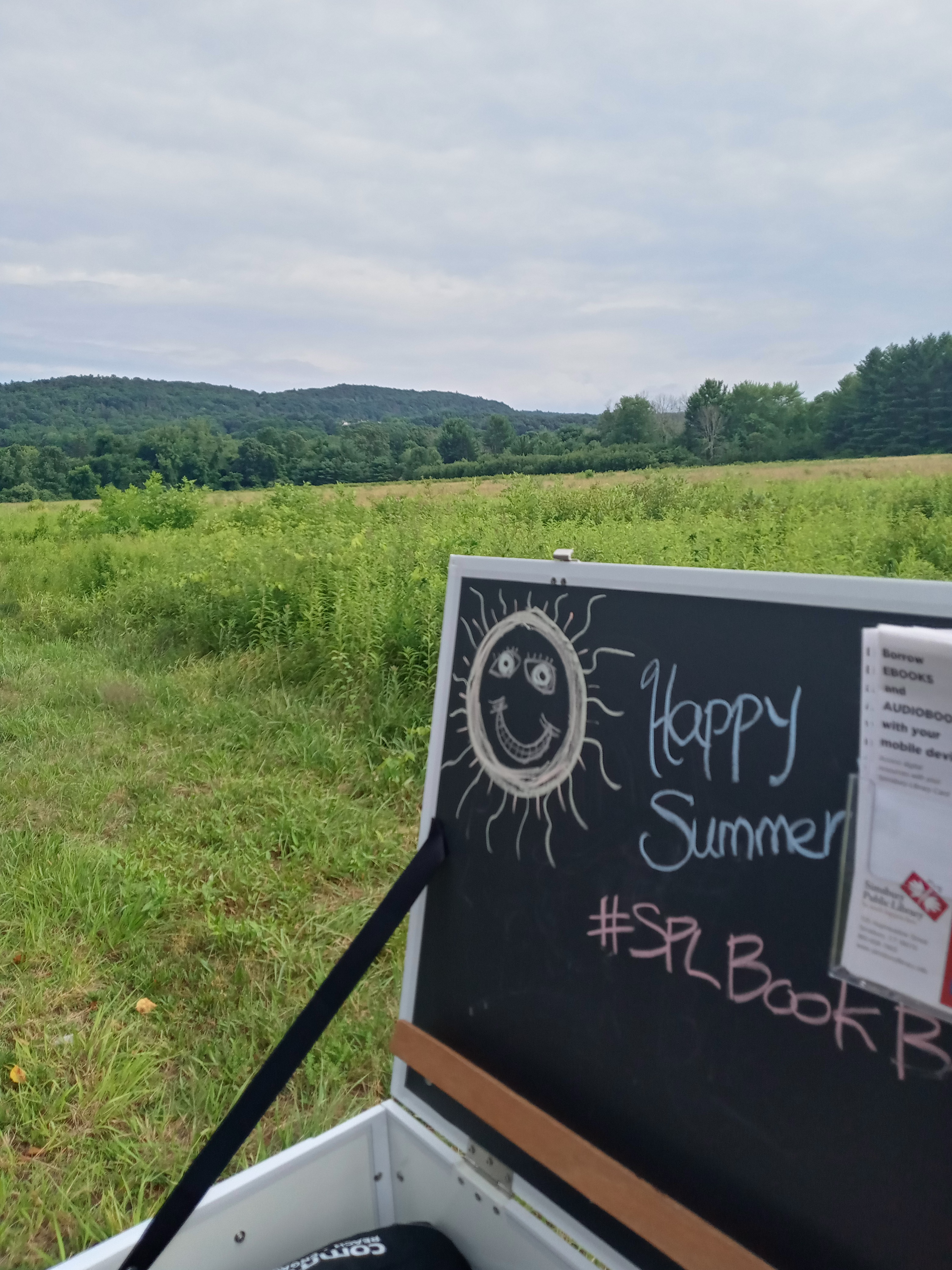 Simsbury Public Library Book Bike with Happy Summer on chalkboard and view of Simsbury fields